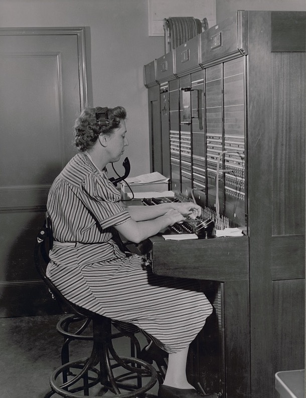 A telephone operator seated at a large switchboard, plugging in a cord, 1941